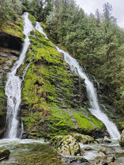 Waterfall cascading down a jagged, moss-covered rock face, surrounded by dense evergreen trees. The water flows into a clear pool at the base, with large, smooth rocks scattered throughout the stream. The sky is overcast, providing a muted backdrop to the lush greenery.