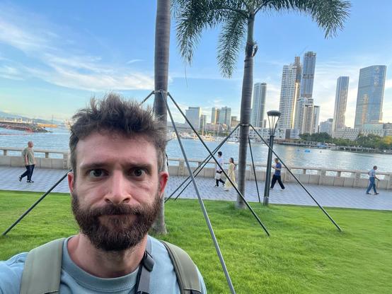A man grimaces towards the camera. Behind him there’s a patch of grass with some trees, a sidewalk with some people, and farther back some tall buildings