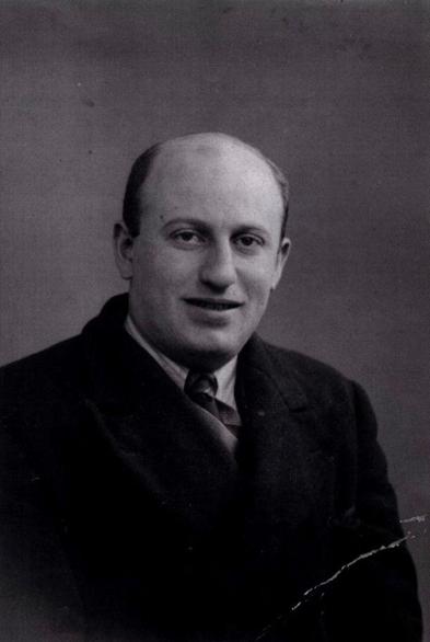 Black and white portrait of a man, sitting and facing the camera. He is wearing a dark suit with a light-colored shirt and a tie. The background is plain and neutral.
