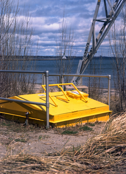 A low to the ground, bright yellow structure at the base of an transmission pylon looks like it may be the entrance to an underground hideout. Lake Ontario and a lighthouse at the entrance to Hamilton Harbour are in the background.