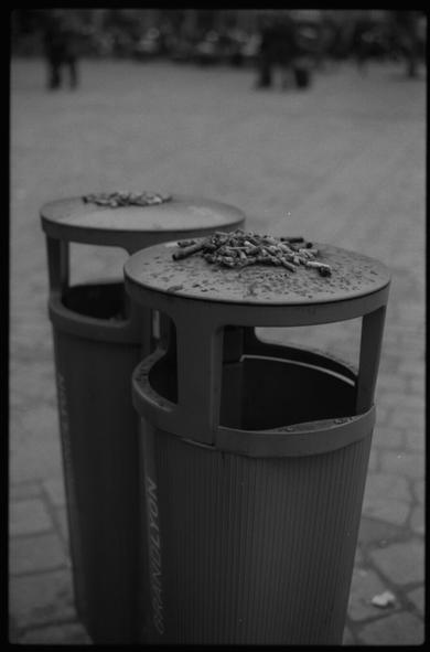 Black and white photography of two waste garbage can in the street. They are both covered with cigarette butts.