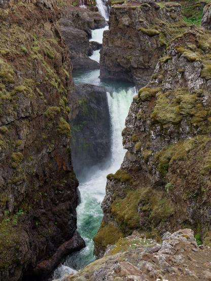 A waterfall cascades between steep rocky cliffs, surrounded by lush greenery. The rushing water creates a vibrant, foamy turquoise pool at the base.