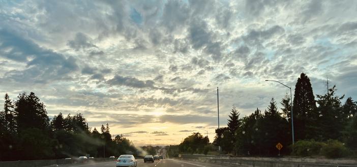 Looking through my windshield at a long road ahead, lined on each side with lots of trees. The sky is filled with billowy clouds. It’s a gradient color, beginning with a light blue gray overhead, continuing to a light gold where it meets the earth. The bright sun is trying to peek through a layer of clouds near the horizon.