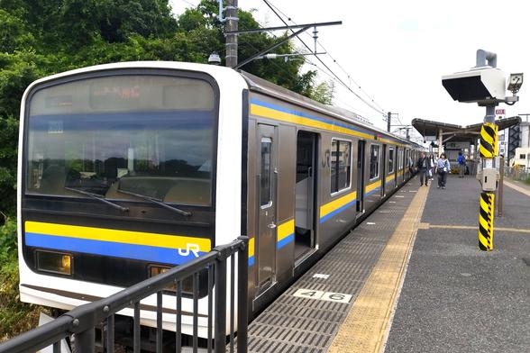 A gray JR commuter train with a thin yellow-and-blue stripe down the side sits at a mostly open-air platform on an overcast day. A bit of the platform (including the CCTV post for the conductor, some passengers) are also in the shot.