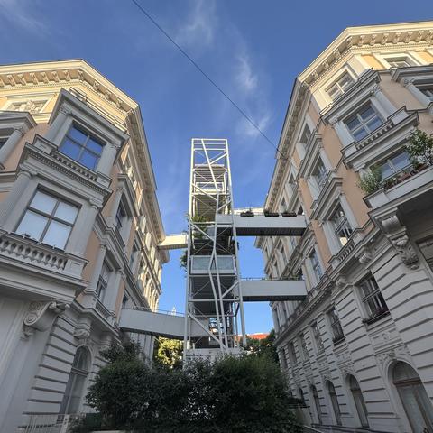 A narrow, elongated metal tower between two massive baroque houses. From a few windows bridges lead to the central metal structure. 
There is a bush in front of the tower and the sky is blue with a few cirrus clouds.