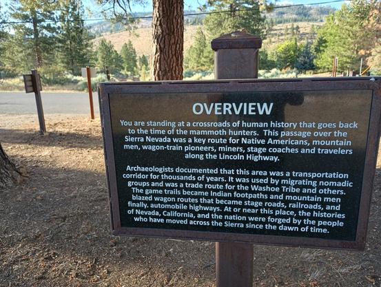 A color landscape photo of a rusted metal framed black sign that reads in white lettering: "Overview. You are standing at a crossroads of human history that goes back to the time of the mammoth hunters. This passage over the Sierra Nevada was a key route for Native Americans, mountain men, wagon-train pioneers, miners, stage coaches and travelers along the Lincoln Highway. Archaeologists documents that this area was a transportation corridor for thousands of years. It was used by migrating nomadic groups and was a trade route for the Washoe Tribe and others. The game trails became Indian footpaths and mountain men blazed wagon routes that became stage roads, railroads, and finally, automobile highways. At or near this place, the histories of Nevada, California, and the nation were forged by the people who have moved across the Sierra since the dawn of time." Conifer trees and a sunlight dry grass field are seen above and to the left of the sign.
