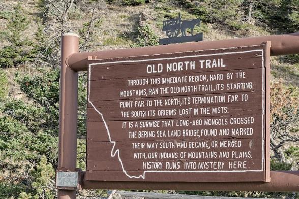 A color landscape photo of a brown wooded sign attached to large vertical and horizontal pipes painted the same color as the sign. The sign has white lettering inscribed in and outline representative of the US state of Montana. The sign reads, "Old North Trail. Through this immediate region, hard by the mountains, ran the Old North Trail, its starting point far to the north, its termination far to the south its origins lost in the mists. It is a surmise that long-ago mongols crossed the Bering sea land bridge, found and marked their way south and became, or merged with, our Indians of mountains, and plains. History runs into mystery here." There's a small black metal cutout of a horse and buckboard on top of the sign. A second small sign at the bottom left reads, "Bruner, Paul - Olvie, BLT 7 1977." A hill with brush is seen behind the sign.