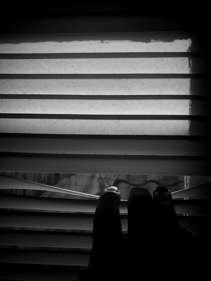 Black and white photo of a detail of white window blinds. In the bottom of the frame three fingers can be seen pulling on eof the slats slightly downward, revealing part of a roof behind the window.