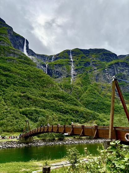 Green mountains with three waterfalls. A wooden bridge in the foreground, crossing a wide river. The sky is overcast.