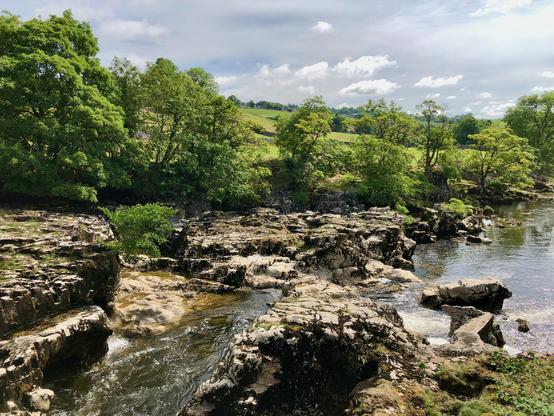 This picture showcases a serene natural landscape with a rocky riverbank and gently flowing water. The river meanders through rugged, uneven rocks, creating small cascades and pools. On both sides of the river, lush green trees and shrubs thrive, indicating a healthy, verdant environment. The background features rolling green hills under a partly cloudy sky, with patches of sunlight breaking through to illuminate the vibrant landscape. The overall scene exudes tranquility and natural beauty, typical of a peaceful countryside setting.