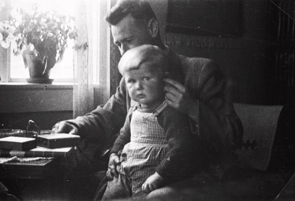 A black and white photo of a toddler on the lap of a man, both listening to a small radio device.