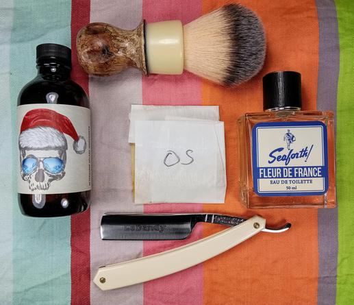 (Le Chat Mistral description)

The image showcases a collection of grooming items arranged on a striped background. At the top left, there's a dark bottle with a label featuring a skull wearing sunglasses and a Santa hat. Next to it, there's a shaving brush with a wooden handle. In the centre, there's a small packet of tissues. To the right, there's a bottle of Seaforth! Fleur de France eau de toilette. At the bottom, there's a straight razor with a cream-coloured handle and the brand "LeDandy" inscribed on the blade. The items are neatly laid out, suggesting a well-organised grooming routine.
