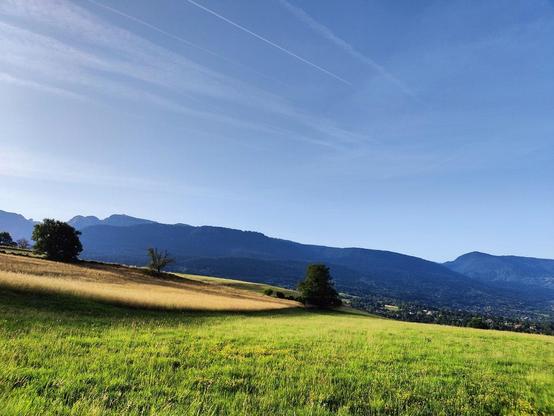 Photo d'un paysage. Un ciel un peu voilé avec quelques traits blancs. Des montagnes en arrière-plan. Puis, plus proches, des collines de prés jaunes, puis verts.