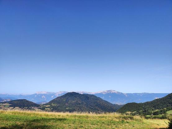 Photo de paysage du massif d'en face, sous un grand ciel bleu, et derrière quelques collines vert foncé.