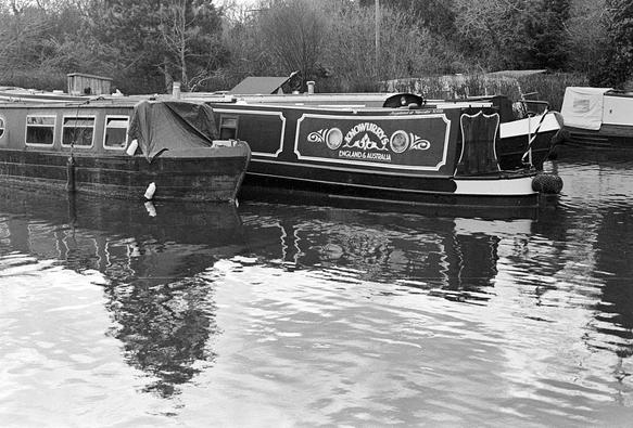 Black and white photo of canal boats (known as narrow boats) moored pointing outwards into a canal basin. The left one has a tarpaulin over its bow area. The middle one is stern out and has a decorative panel on the side that reads "KNOWURRYS ENGLAND & AUSTRALIA". The boats behind cannot be seen clearly. There are winter trees behind, some reflected in the slightly rippled water in the foreground.