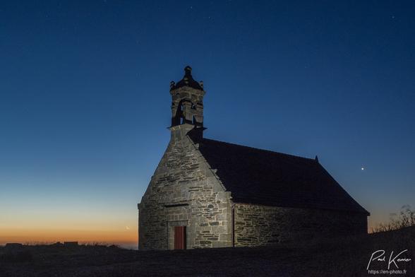 Photographie en couleur prise de nuit sous un ciel clair, étoilé, d'une chapelle au sommet d'une montagne. 
Au pied de la façade de la chapelle, un léger éclairage de leds pallie au contre-jour de fin de nuit, alors qu'à l'arrière-plan, au nord-est, le soleil se lèvera dans quelques dizaine de minutes, et déjà l'horizon est bien coloré en orange avec au-dessus, une bande bleuâtre qui pourrait être la lueur des nuages noctulescents présents plus au nord. 
Juste à gauche de la chapelle, on peut voir un fin croissant.(2%) de lune, qui vient de se lever, alors qu'à droite de la chapelle, un peu plus haut dans le ciel, on distingue nettement Vénus, l'étoile du berger. 
Un petit clocher domine la façade de la chapelle où on peut voir une porte en son centre.