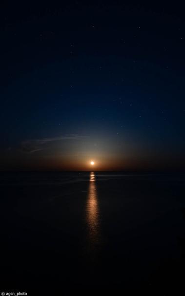Blick auf das dunkle Meer mit dem aufgehenden Vollmond, der sich im Wasser spiegelt. /
View of the dark sea with a rising full moon reflecting on the water.