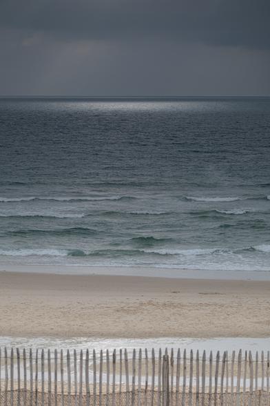 Photo de la plage de Carcans sous un ciel chargé. Elle montra au premier plan une barrière surplombant une plage, puis au loin il y a l'océan et le ciel grisonnant. On perçoit à l'horizon le reflet des rayons qui percent les nuages.