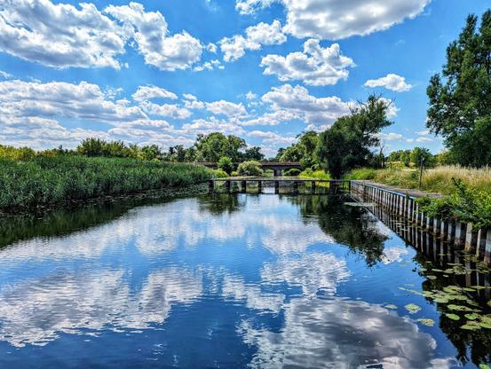 A picturesque natural landscape shows a calm, dark blue river reflecting a bright blue sky dotted with fluffy white clouds. Lush green reeds and trees line the left bank, while the right bank features tall grass and what appears to be a worn wooden embankment with some metal posts. In the mid-ground, a small, multi-arched stone bridge spans the river, partially obscured by the surrounding vegetation. The overall impression is one of a tranquil, sunny day in the countryside.
