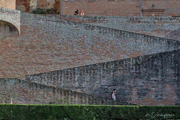 A green hedge topped by a pink and grey brick wall. A staircase runs diagonally down the wall. At the very top, a couple are leaning against the wall. At the bottom, level with the hedge, another couple is descending.