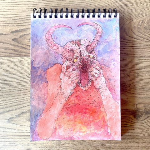 Fotografía de un cuaderno con un dibujo de una criatura homanoide con grandes cuernos una boca feroz abierta por ambas manos. La técnica es acuarela y tinta, la acuarela son manchones de colores amarillos y rojos para la criatura y azul para el fondo