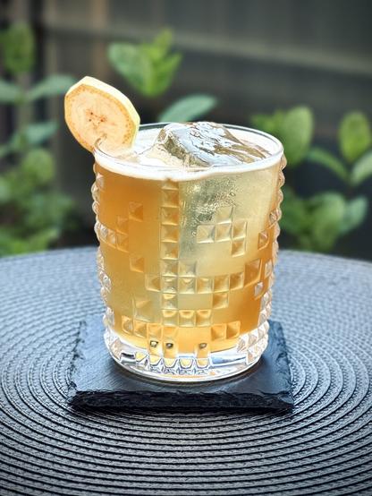 a golden yellow cocktail with a large rock of clear ice and banana slice garnish sitting on a black slate coaster with mint in the background