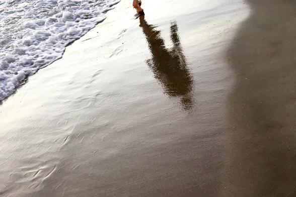 Der Schatten einer gehenden Person am Sandstrand. Ganz oben im Bild sieht man noch den Fuß der Person, ansonsten nur ihren fast senkrechten Schatten nach unten. Sie geht am Meeressaum, links im Bild reichen die Wellenausläufer fast an den Fuß heran. Der Boden ist leicht feucht, ganz schwach erkennt man noch die Fußspuren. Sie wurde größtenteils schon vom Meer verwaschen.