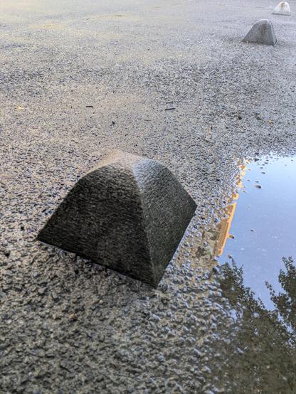 Three pyramidal concrete teeny tiny bollards on a university path retreat into the distance. A puddle to the right reflects blue sky and clouds and just the edge of the science building.