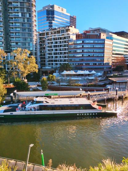 A closer look at the ferry, looking downward from across the river. 

The bottom half of the ferry is dark green with the word "RiverCat" in white letters, and the top half is white. It has outdoor seating at the front and rear. 

Behind it are trees, tall office buildings, and the outdoor dining areas of restaurants with tables under white umbrellas.