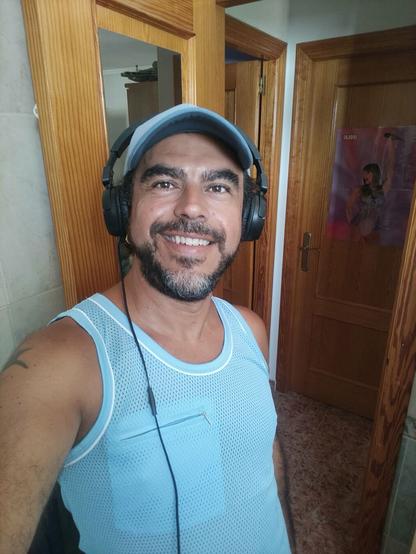 Un homme souriant porte un débardeur bleu clair et un casque audio noir. Il porte une casquette bleue et a une barbe. Il est debout dans une salle de bain avec des portes en bois visibles en arrière-plan.  Sur une porte, un poster d'une chanteuse est affiché. L'homme semble détendu et à l'aise.