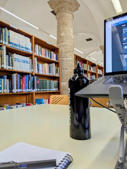 Foto desde mi puesto de trabajo de unas estanterías de la biblioteca con uno de los viejos pilares del antiguo hospital en el centro, y mi portátil, libreta, pluma y botella de agua en primer plano