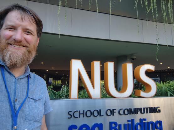 A bearded man stands next to a sign that reads 'NUS School of Computing'. Some tropical plants can be seen behind / above it.