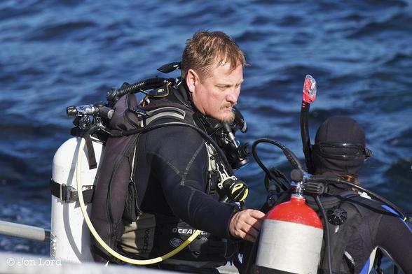 A colour waterside photo looking downwards at two divers on a platform preparing to enter the water. In the centre is a young diving instructor, In profile and facing to the right. He has short fair hair and goaty beard: his face lit by the still low morning sun. He is wearing a black wetsuit and has an air tank on his back with the mask over his left shoulder. The instructor is checking The Equipment of his female student diver who to the right with her back to the camera. She is dressed and equipped as the instructor except she has a snorkel with a pink day-glow top attached to her head. In the background is a dark blue and choppy pacific ocean.
The location: Santa Catalina, California (2014).