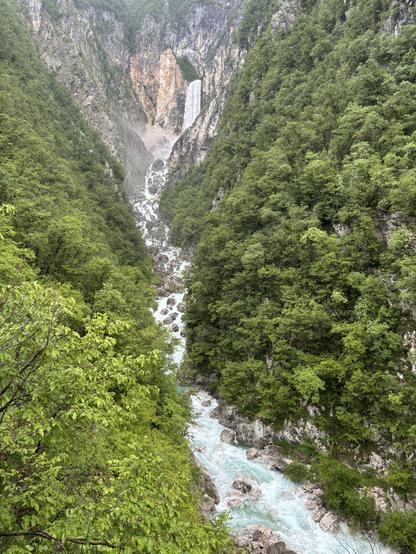 A waterfall seen from a distance. The water runs through a steep, verdant mountain gorge.
