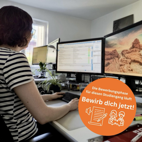 Das Bild zeigt eine junge Frau mit kurzen dunklen Haaren, die an einem Schreibtisch vor zwei großen Computerbildschirmen sitzt und arbeitet.