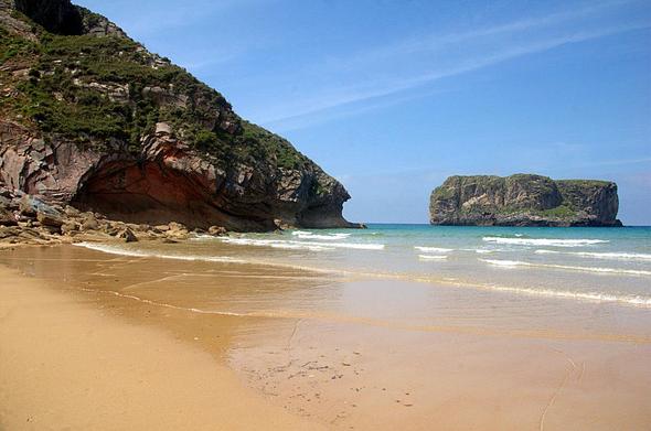 Playa bajo un acantilado de rocas pelirrojas que se rizan divertidas, la mar tranquila relame con leves olas la húmeda arena que parece turrón del blando. Al fondo un pequeño islote se baña apacible los bajos. Es una conocida playa de Llanes por ser escenario de varias películas, aquí sin focos ni directores ni actores ni espectadores, desnuda.