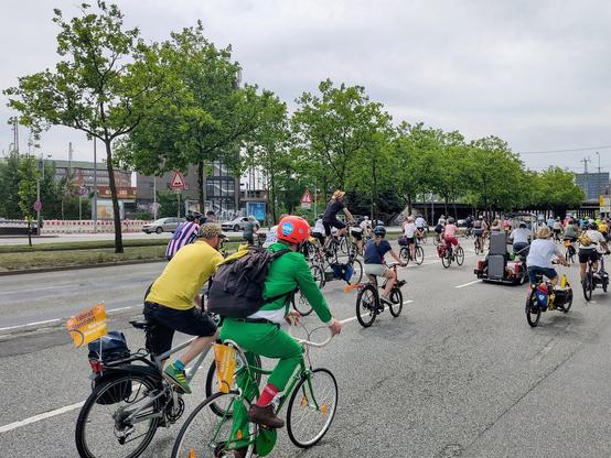 Viele Fahrradfahrer:innen auf einer Straße mit grünen Bäumen auf dem Mittelstreifen. Im Vordergrund ein Fahrradfahrer mit komplett grünem Anzug auf einem schicken grünen Fahrrad.