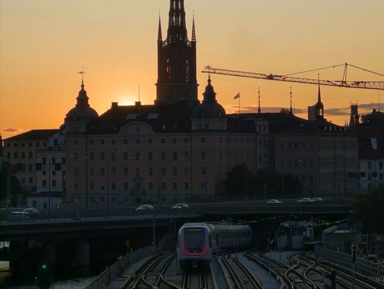 A modern metro train driving along a section with six tracks into a tunnel under an elevated road, behind which the sun is setting behind some old European buildings