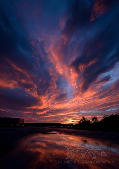 A dramatic sunset fills the sky with vibrant orange, red, and dark blue clouds, which are reflected in a large puddle on a wet asphalt surface in the foreground. Silhouetted trees and what appears to be a building are visible on the horizon.