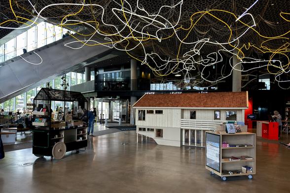 A modern indoor space features a large model of a house with a wooden exterior and a red roof. In the foreground, there is a display cart with books and items for sale. The ceiling is adorned with artistic lighting fixtures, and large windows allow natural light.