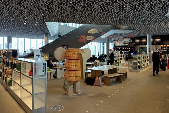 A modern library interior featuring wooden shelving filled with books, a unique elephant-shaped seating structure, and a spacious design with an intricate ceiling pattern. People are seen interacting in the space, with a staircase in the background and decorative elements hanging from the ceiling.