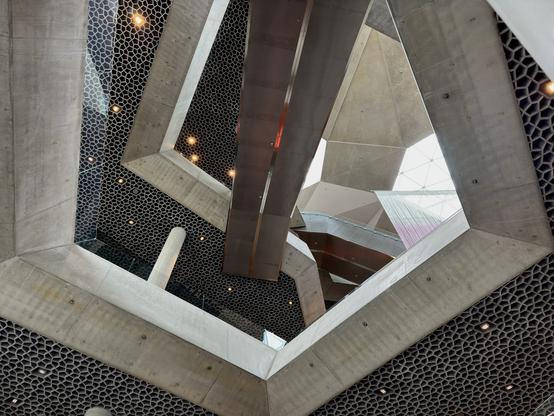 An interior view of a modern architectural space featuring a geometric ceiling with a honeycomb pattern, a skylight, and suspended light installations resembling abstract shapes. The design incorporates concrete elements and glass, with visible signage and furniture below.