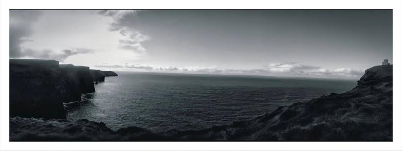 The photo is a panoramic, black and white shot of the Cliffs of Moher in Ireland.  The vast Atlantic Ocean is in the centre and stretches out to a hazy horizon under a mostly cloudy sky.  The rugged cliffs, on top covered in grass, frame the scene on both sides, with  O'Brien's tower on the right-hand cliff.
