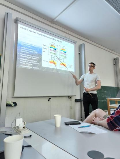 A nerdy-looking man points a stick at projector screen showing a slide describing species distribution modelling.