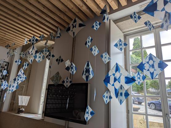 Inside a room with wooden beams on the ceiling, from which are hanging many blue and white paper diamonds, which are actually cyanotype pictures of origami cranes. The bottom border of the hanging has the same form as the river in this place, Ponte de Lima