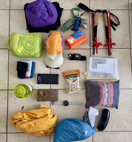 Flat lay of neatly arranged hiking gear on a tiled floor, including clothing, trekking poles, rope, water bottle, snacks, headlamp, cap, sunglasses case, solar power bank, and other essentials for a tour from hut to hut.