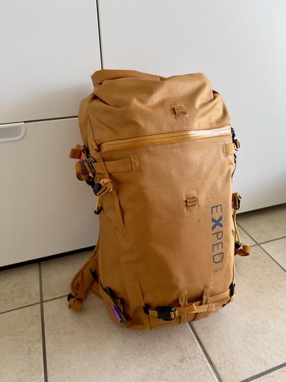 A large orange EXPED hiking backpack sits upright on a tiled floor, packed and ready for an outdoor adventure. The backpack features multiple straps, buckles, and a front zipper pocket, designed for durability and functionality.