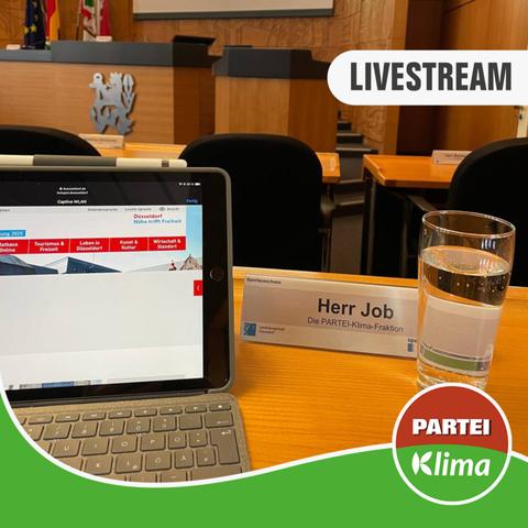 Zu sehen ist eine Nahaufnahme des Tisches von Stefan Job (Klimaliste) im Plenarsaal des Düsseldorfer Rathauses. Im Fokus ist der geöffnete Laptop und ein Glas Wasser. Im Hintergrund ist das Redepult und der Sitz des Ausschussvorsitzenden.