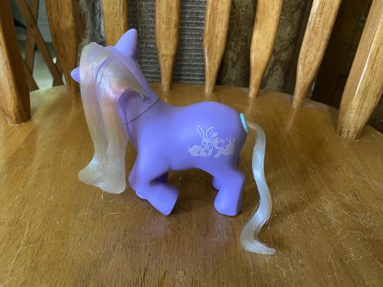 A small purple toy pony with long, shiny hair, displayed on a wooden surface. It features a playful design of rabbits on its side.