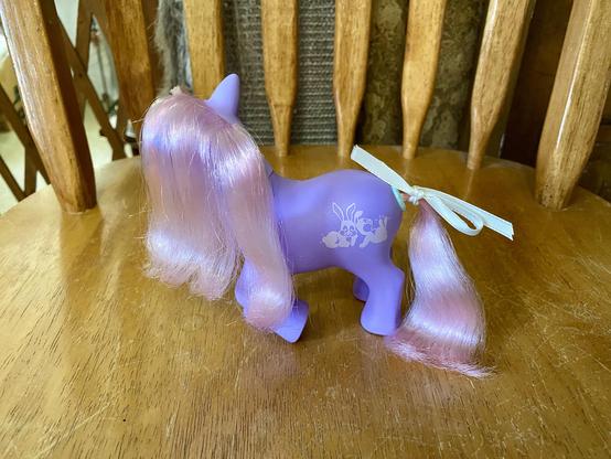A purple toy pony with long, shiny pink hair and a ribbon tied in its tail. The pony features a design of small cartoon bunnies on its side. The figure is positioned on a wooden table.